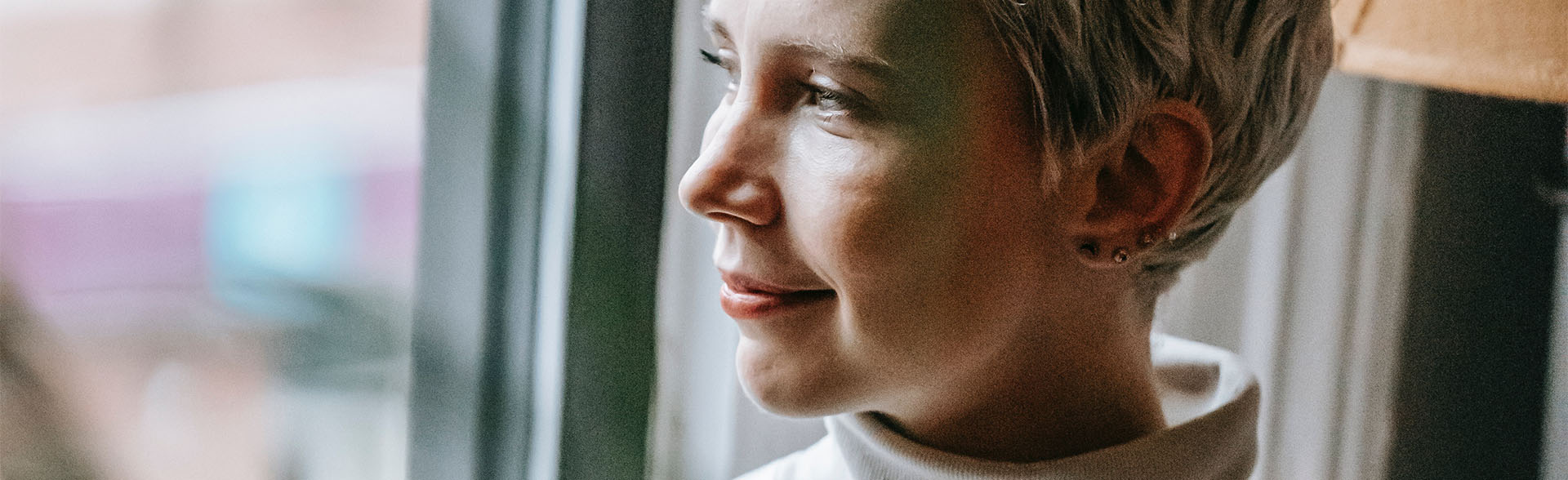 Frau mit kurzen, weißen Haaren und weißem Rollkragenpullover blickt lächelnd aus dem Fenster.